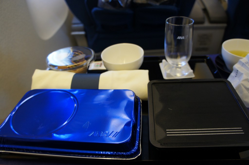 The image shows a meal setup on an airplane tray table. The setup includes a blue foil-covered meal tray, a black tray, a white napkin, a glass with the ANA logo, a small bowl, and a cup with a beverage. The background shows part of the airplane seat and window.