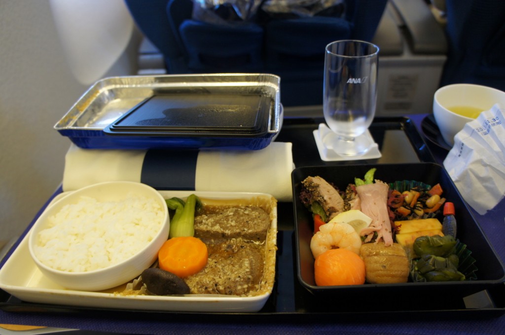 The image shows an in-flight meal served on an airplane tray table. The meal includes a bowl of white rice, a dish with a piece of meat in sauce, a carrot slice, a piece of mushroom, and some green vegetables. There is also a bento-style box containing various items such as shrimp, a slice of ham, a piece of fish, assorted vegetables, and a small bottle of sauce. Additionally, there is an empty glass with the airline's logo, a cup of tea, and a napkin on the tray. The background shows airplane seats.