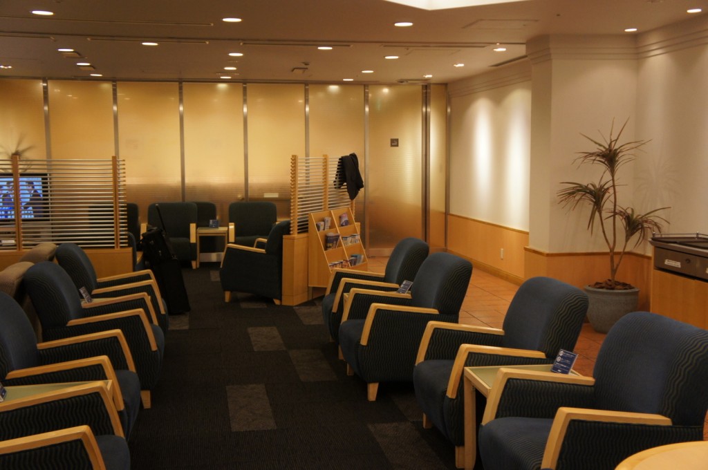 The image shows a waiting area or lounge with several blue cushioned chairs arranged in rows. The chairs have light wooden armrests. There is a small table with brochures or magazines in the center of the room. The walls are light-colored, and there is a potted plant in the corner. The lighting is warm, and the room has a calm and organized atmosphere. There is also a frosted glass partition in the background, separating this area from another space.