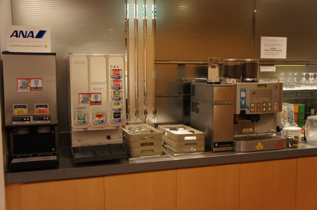 The image shows a beverage station in what appears to be a lounge or cafeteria. The station includes three main machines: a hot beverage dispenser on the left, a Coca-Cola branded soda fountain in the middle, and a coffee machine on the right. There are trays with cups and lids in front of the machines, and a sign with the ANA logo (All Nippon Airways) is visible above the hot beverage dispenser. The background features a metallic wall with a shelf holding glasses and other items.