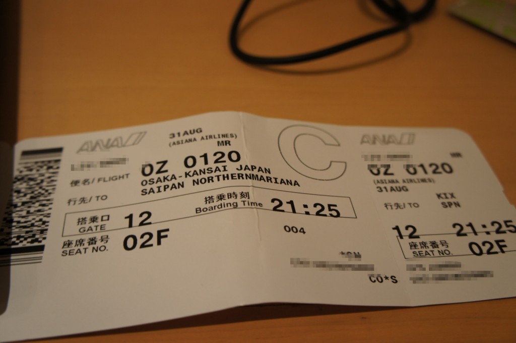 The image shows a boarding pass for a flight. The details on the boarding pass include:
- Airline: Asiana Airlines (ANA)
- Flight number: OZ 0120
- Date: 31st August
- Departure: Osaka-Kansai, Japan (KIX)
- Destination: Saipan, Northern Mariana Islands (SPN)
- Gate: 12
- Boarding time: 21:25
- Seat number: 02F
- Class: C
The boarding pass is placed on a wooden surface, and there is a black cable visible in the background. Some personal information on the boarding pass is blurred out for privacy.