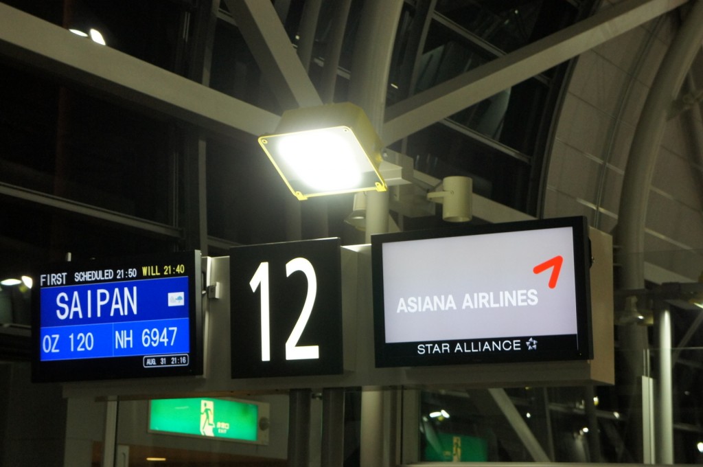 The image shows an airport gate sign. The gate number is 12. There are two digital screens: one on the left displaying flight information and the other on the right showing the Asiana Airlines logo with the text "ASIANA AIRLINES" and "STAR ALLIANCE." The flight information screen indicates a flight to Saipan with flight numbers OZ 120 and NH 6947. The scheduled departure time is 21:50, and the current time is 21:16. An emergency exit sign is visible in the background.