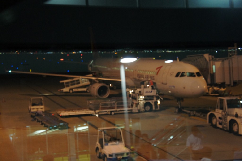 The image shows an airplane parked at an airport gate at night. The aircraft is connected to a jet bridge, and there are various ground service vehicles around it, including a baggage cart and a pushback tug. The airport runway and taxiway lights are visible in the background, along with the city lights in the distance. The photo appears to be taken from inside the airport terminal through a window.