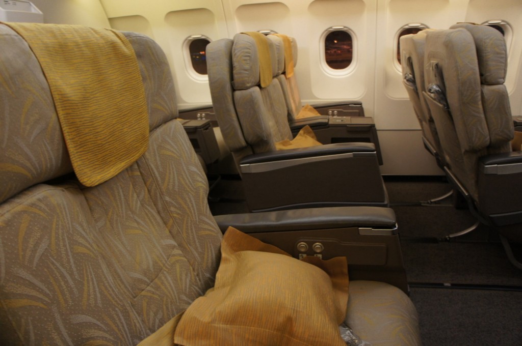 The image shows the interior of an airplane cabin, specifically the business or first-class section. The seats are wide and upholstered in a patterned fabric with a yellow headrest cover. Each seat has a yellow pillow placed on it. The seats are arranged in pairs, with ample legroom and individual armrests that include control buttons. The cabin has windows along the side, and the lighting is warm and soft.