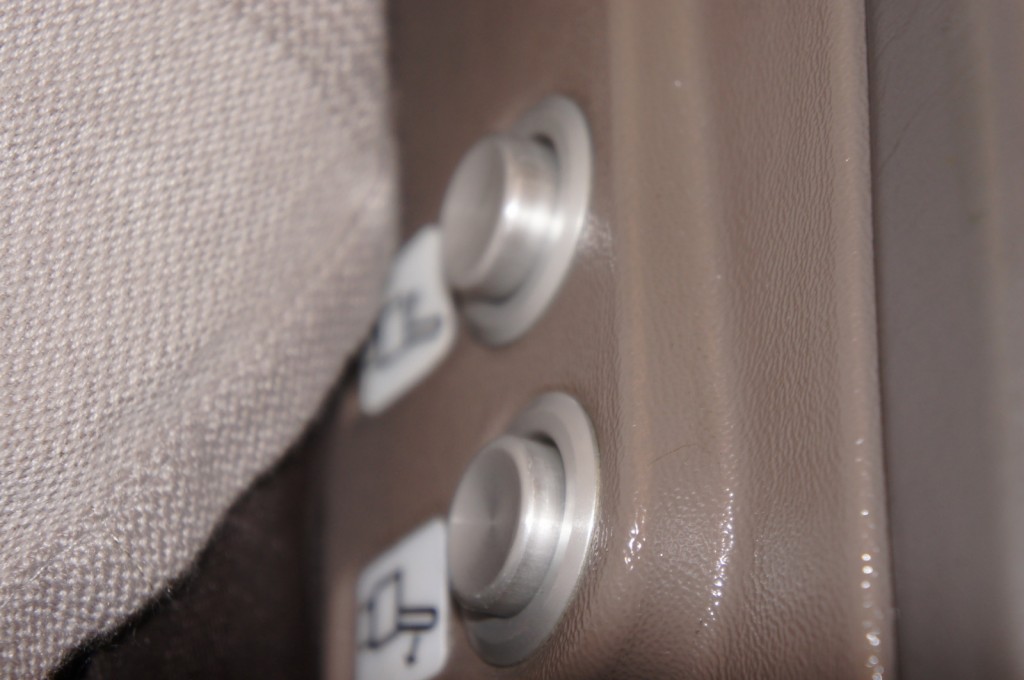 The image shows a close-up view of two metallic buttons on a textured surface, likely part of a seat or armrest. The buttons have small icons next to them, which appear to be related to seat adjustments. The background includes a fabric material, possibly part of the seat upholstery.