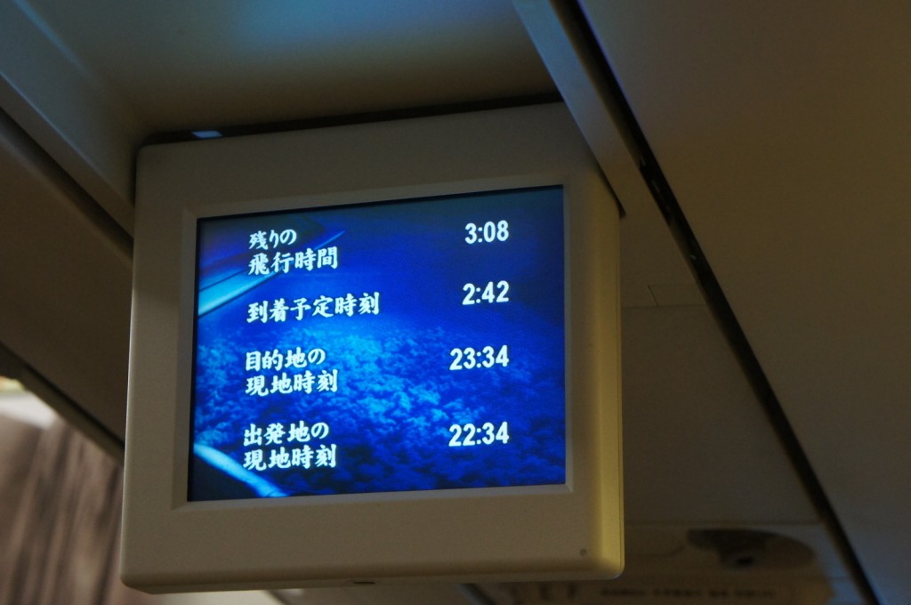 The image shows an in-flight screen displaying flight information in Japanese. The screen is mounted on the ceiling of an airplane. The information includes the remaining flight time (3:08), the estimated arrival time (2:42), the current time at the destination (23:34), and the current time at the departure location (22:34). The background of the screen shows an image of clouds.