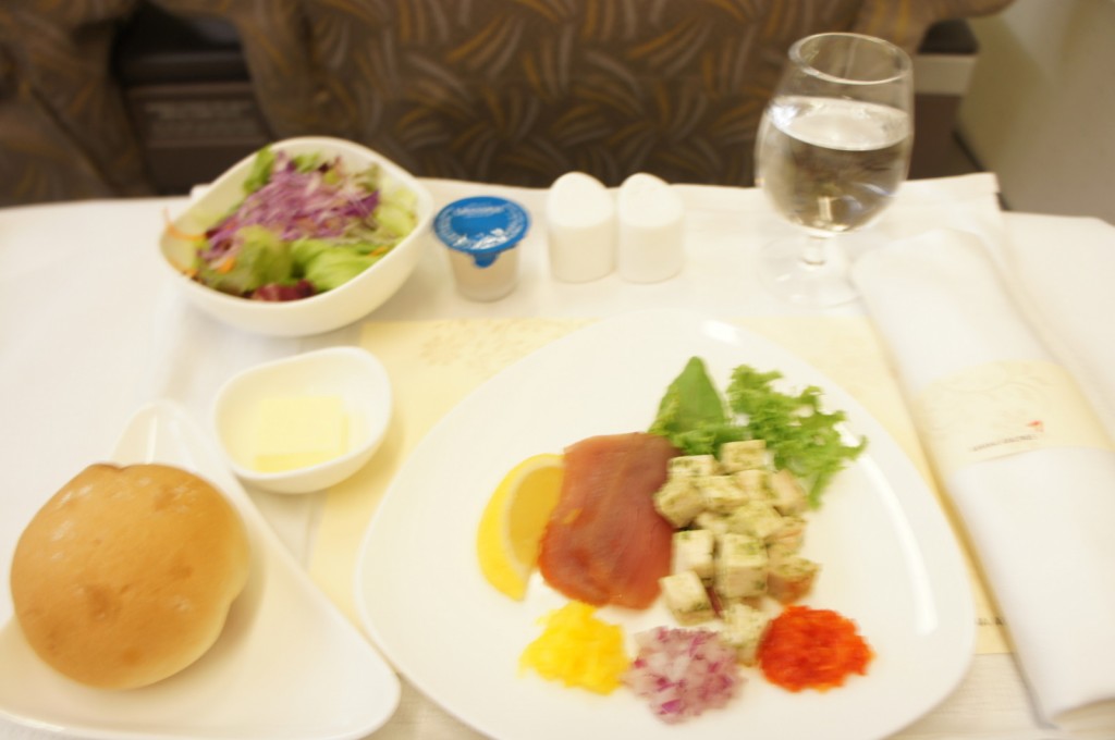 The image shows a meal served on a tray, likely on an airplane. The meal includes a plate with a variety of foods such as a slice of lemon, diced vegetables, a piece of lettuce, and what appears to be a slice of meat or fish. There is also a small bowl of butter, a bread roll, a bowl of salad with mixed greens and shredded purple cabbage, a small container of dressing or sauce, and a glass of water. Additionally, there are two small containers, possibly for salt and pepper, and a rolled-up napkin with a napkin ring.