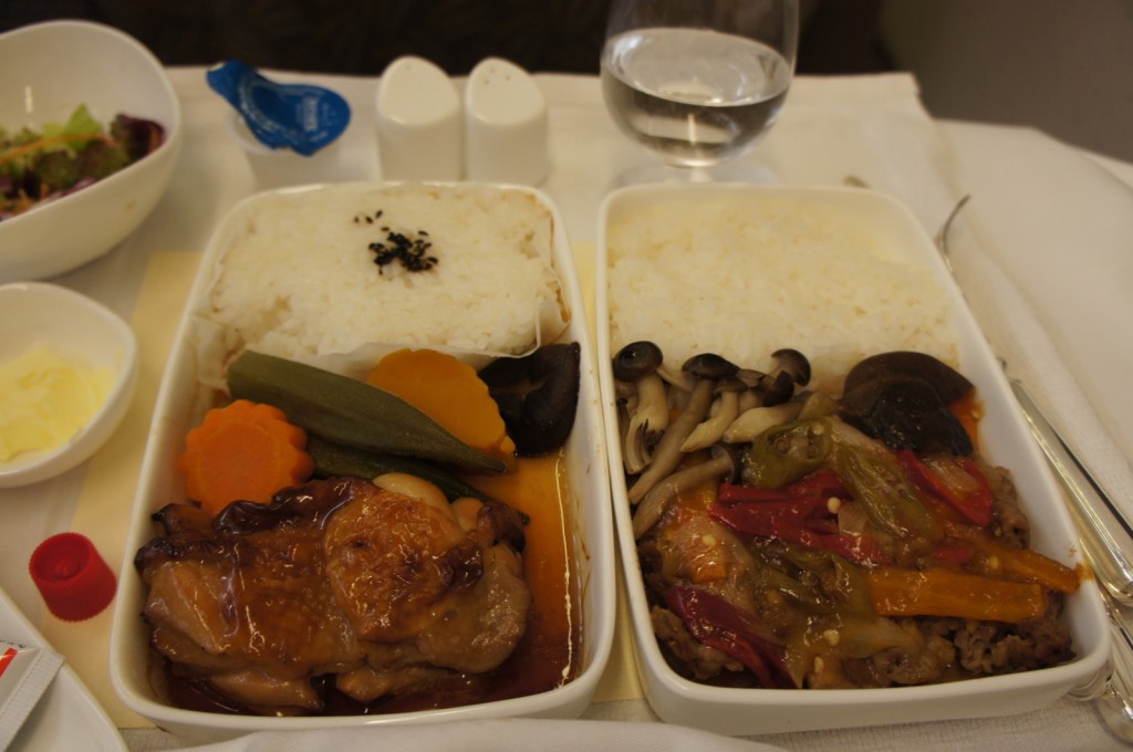 The image shows a meal served on a tray, likely on an airplane. There are two main dishes in rectangular containers. The left container has white rice topped with black sesame seeds, a piece of glazed chicken, and a variety of vegetables including carrots and green beans. The right container also has white rice and a mix of vegetables including mushrooms and bell peppers. Additionally, there is a small bowl of salad, a small dish of butter, a glass of water, and two small condiment containers on the tray.