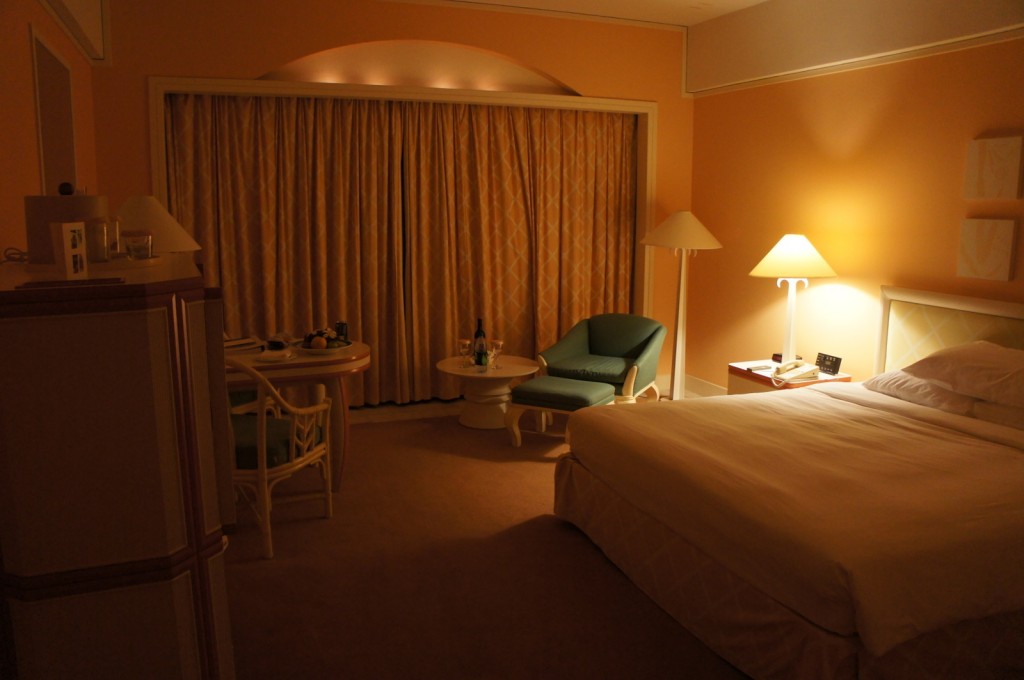 A warmly lit hotel room with a large bed on the right side, covered in white linens. Next to the bed is a nightstand with a lamp, a telephone, and an alarm clock. In the center of the room, there is a small round table with two chairs, and a bottle of water and some glasses on the table. To the left of the table is a green armchair with an ottoman. The room has beige walls and a large window covered with heavy curtains. There is also a floor lamp near the armchair and a cabinet on the left side of the image.
