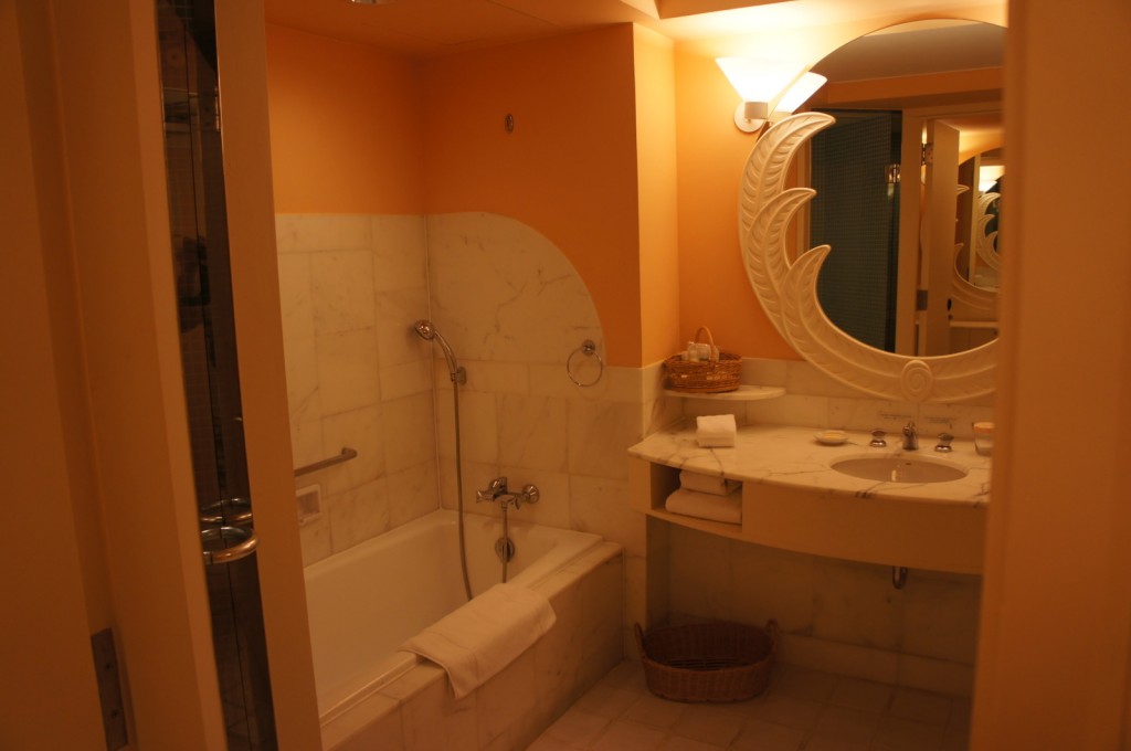 The image shows a bathroom with warm lighting and a cozy ambiance. The bathroom features a bathtub with a handheld showerhead, surrounded by white marble tiles. There is a white towel draped over the edge of the bathtub. Adjacent to the bathtub is a vanity area with a marble countertop, a sink, and a large decorative mirror with a leaf-like design. On the countertop, there are neatly folded towels, a basket with toiletries, and some small items. Below the countertop, there are additional towels and a wicker basket. The walls are painted in a soft peach color, complementing the overall warm and inviting atmosphere of the bathroom.