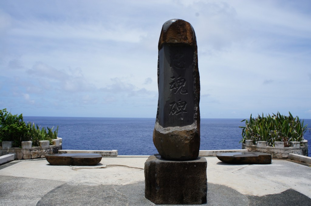 A tall stone monument with engraved characters stands on a concrete platform overlooking the ocean. The sky is partly cloudy, and there are green plants in stone planters on either side of the monument. The scene is serene, with the blue sea and sky in the background.
