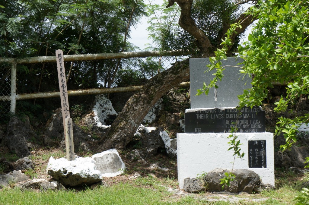 The image shows a memorial site with a stone monument and a wooden post. The stone monument has inscriptions in both English and Japanese. The English inscription reads, "In honor of those who lost their lives during WW-II by Shuichiro Iizuka (Japanese Navy) during 1978 in Saipan." The wooden post also has Japanese writing on it. The memorial is surrounded by greenery, including trees and bushes, and there is a fence in the background. The ground around the monument is covered with rocks and grass.