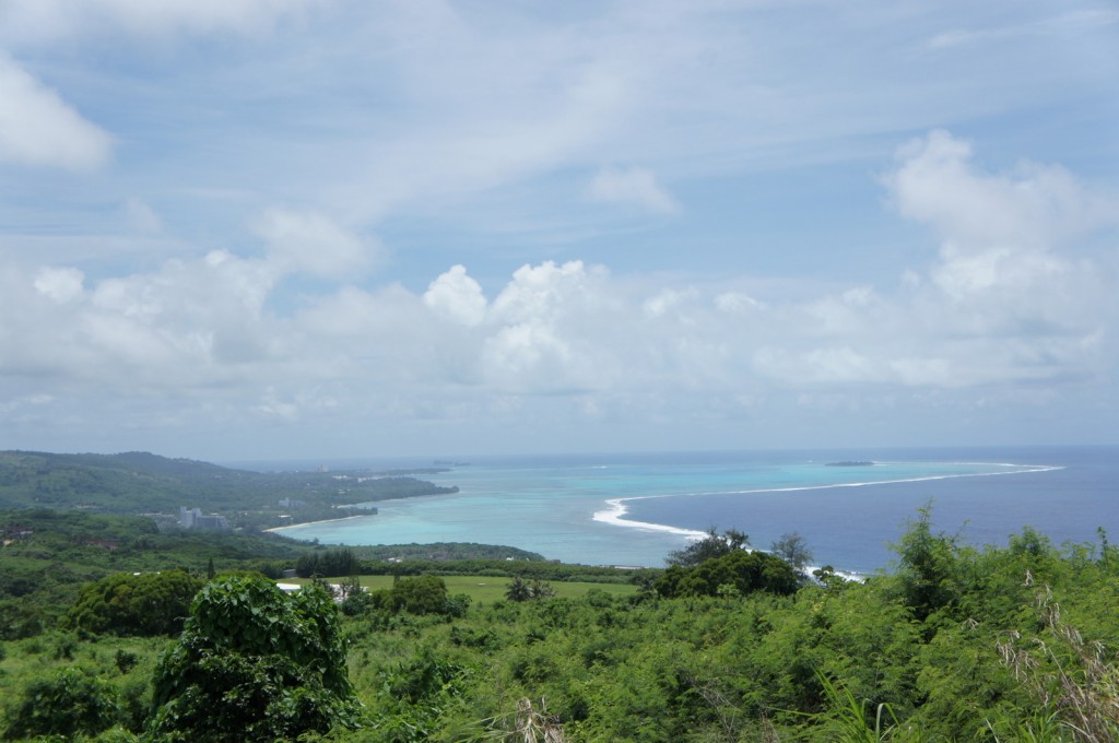 The image shows a scenic view of a coastal landscape. In the foreground, there is lush green vegetation and trees. Beyond the greenery, there is a stretch of land with some buildings visible. The coastline features a beautiful turquoise sea with a white sandy beach and a narrow strip of land extending into the water. The sky above is mostly clear with some scattered clouds, creating a serene and picturesque scene.