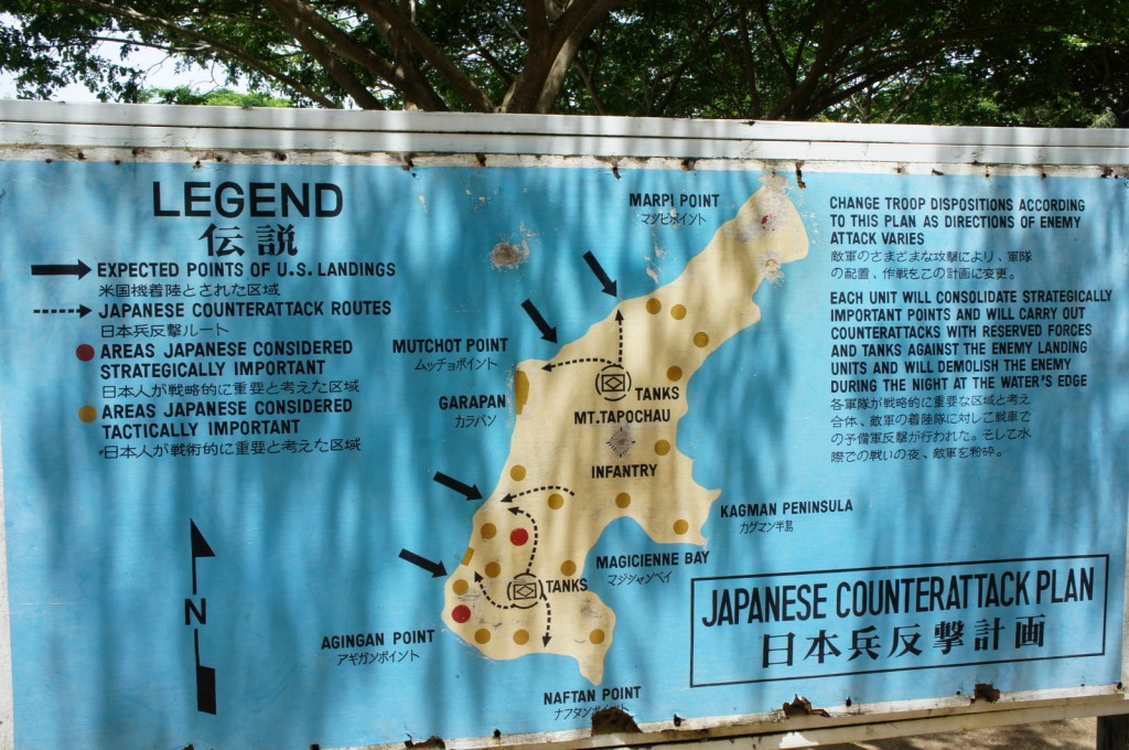 The image shows a detailed map titled "Japanese Counterattack Plan" with both English and Japanese text. The map includes a legend explaining various symbols: expected points of U.S. landings, Japanese counterattack routes, areas considered strategically important, and areas considered tactically important. Key locations such as Marpi Point, Mutchot Point, Garapan, Mt. Tapochau, Kagman Peninsula, Agingan Point, and Magicienne Bay are marked. The map also includes arrows indicating the direction of troop movements and counterattacks. The text on the right side of the map explains the strategy for changing troop dispositions and consolidating important points to carry out counterattacks against enemy landings. The background of the map is blue, and it is displayed outdoors with trees visible in the background.