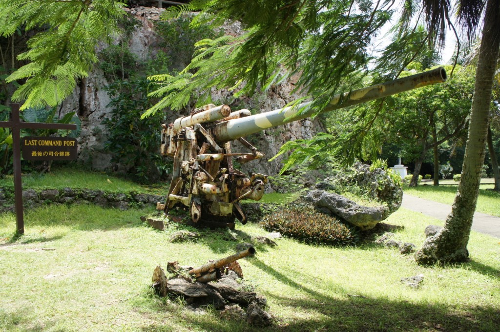 The image shows an old, rusted artillery gun situated in a lush, green outdoor setting. The gun is surrounded by trees and vegetation, with a rocky cliff in the background. To the left of the gun, there is a sign that reads "LAST COMMAND POST" in English and Japanese. The area appears to be a historical site or memorial.