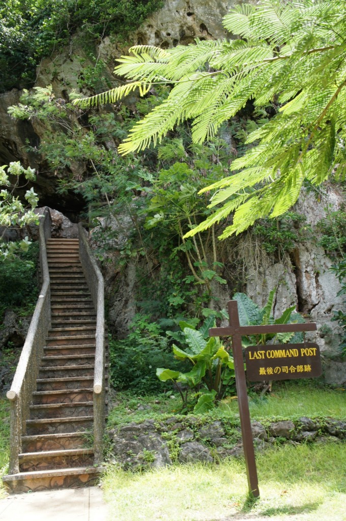 The image shows a stone staircase leading up to a cave entrance surrounded by lush greenery. To the right of the staircase, there is a wooden sign with the text "LAST COMMAND POST" written in English and Japanese. The area appears to be a historical or tourist site, with dense vegetation and rocky terrain.