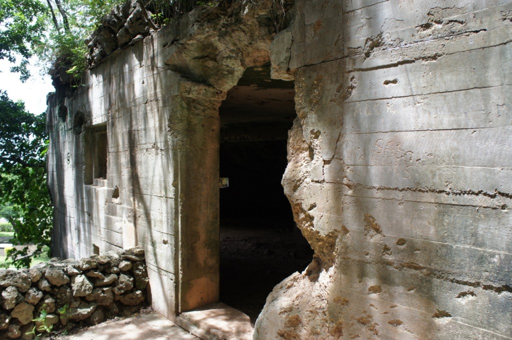 The image shows the entrance to an old, weathered concrete bunker or fortification. The structure has visible damage, with a large hole in the wall near the entrance. The concrete is cracked and eroded, and there are some small windows or openings on the side. The surrounding area is lush with greenery, including trees and plants, and there is a small stone wall in front of the bunker. Sunlight filters through the trees, casting shadows on the structure.