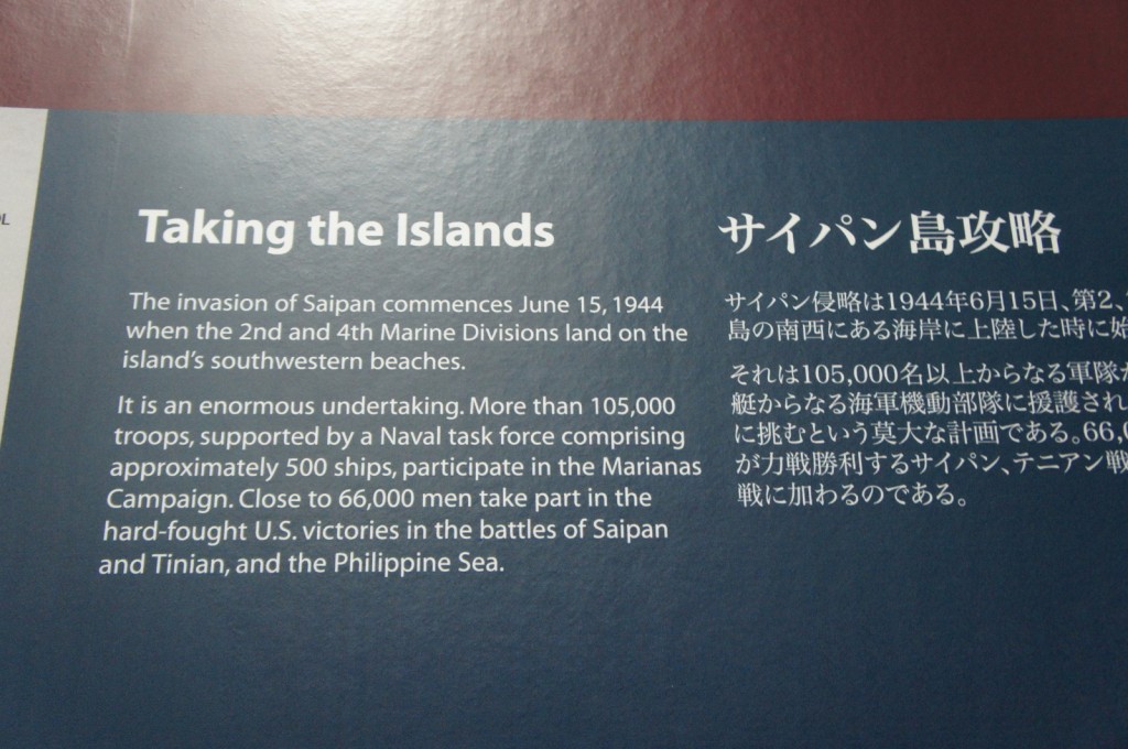 The image shows a display panel with text in both English and Japanese. The English text is titled "Taking the Islands" and describes the invasion of Saipan, which commenced on June 15, 1944, when the 2nd and 4th Marine Divisions landed on the island's southwestern beaches. It mentions that more than 105,000 troops, supported by a Naval task force comprising approximately 500 ships, participated in the Marianas Campaign. Close to 66,000 men took part in the hard-fought U.S. victories in the battles of Saipan and Tinian, and the Philippine Sea. The Japanese text appears to be a translation of the English text.
