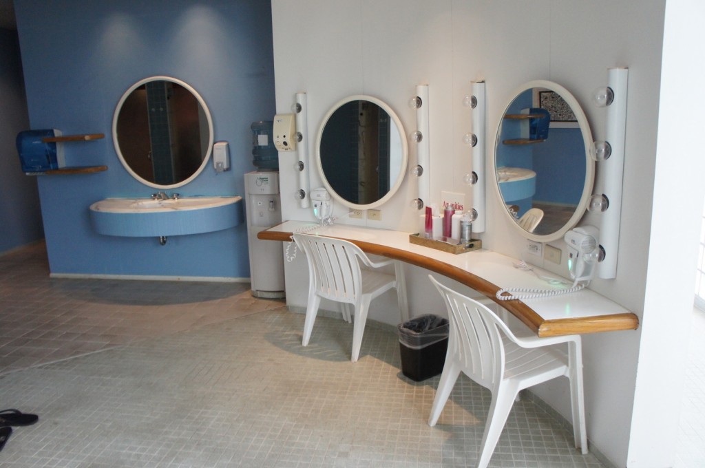 The image shows a well-lit vanity area with two white plastic chairs in front of a curved countertop. Each station has a large round mirror surrounded by light bulbs, resembling a dressing room setup. On the countertop, there are various items including a hairdryer, a small trash bin, and some beauty products. To the left, there is a blue wall with a round mirror above a sink, and a paper towel dispenser mounted on the wall. The floor is tiled, and the overall space appears clean and organized.