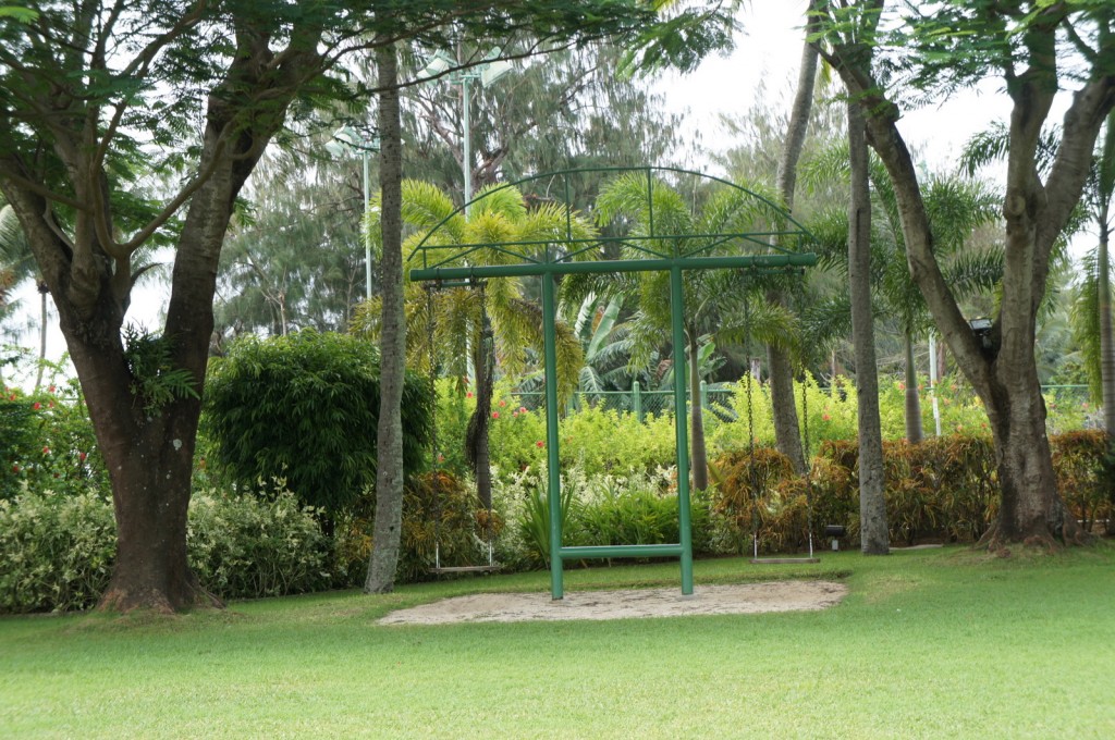 A green metal swing set with two swings is situated in a lush, green park. The swing set is surrounded by various trees and bushes, with a well-maintained grassy area in the foreground. The background features more greenery, including tall trees and dense foliage, creating a serene and natural environment.