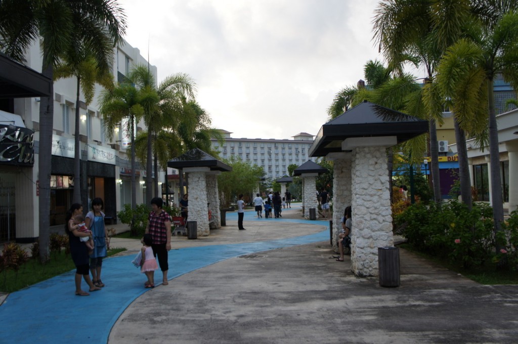 The image shows an outdoor walkway lined with palm trees and stone pillars with black roofs. There are several people walking and standing along the path, including a group of three women and a child on the left side. The walkway has a blue section and a concrete section. In the background, there are buildings with multiple floors, and the sky appears to be overcast. The area has a relaxed, park-like atmosphere with greenery and benches.