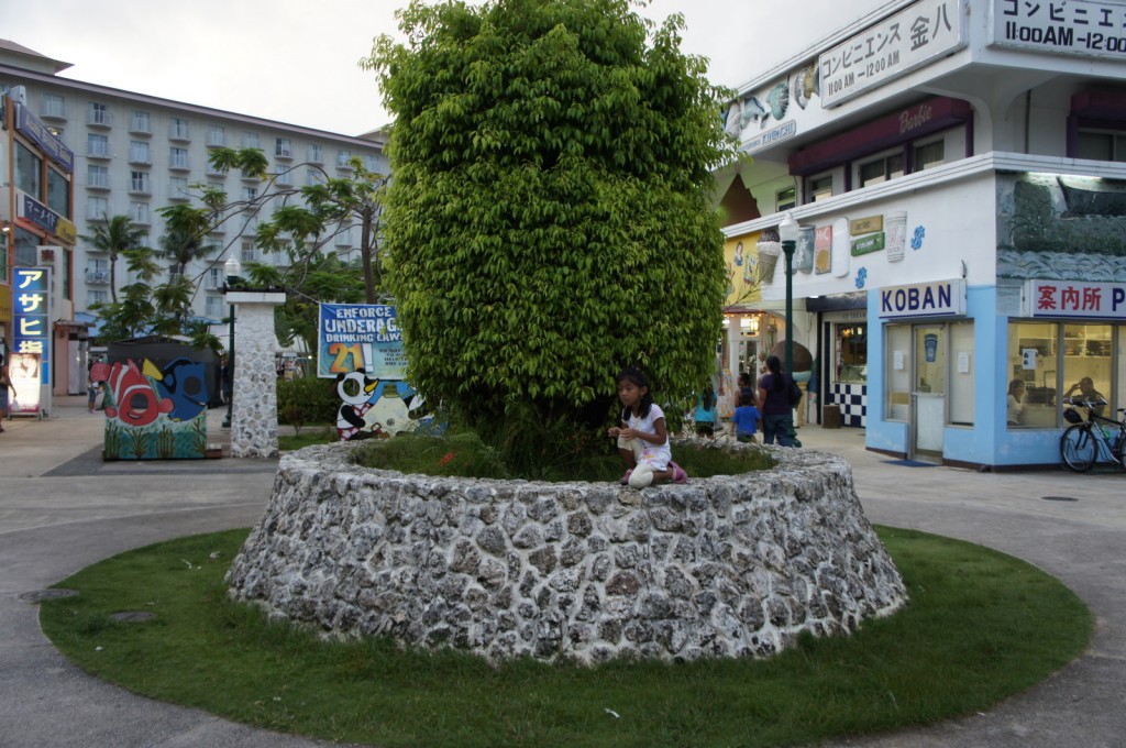 A young girl is sitting on a stone planter with a large, lush green bush in the center. The scene is set in a public area with various buildings and shops in the background. One building has a sign that reads "KOBAN," and another has a sign with Japanese characters. There are also colorful murals and signs, including one that mentions enforcing underage drinking laws. Several people are walking around the area, and there is a bicycle parked near one of the buildings. The overall atmosphere appears to be that of a small commercial or recreational area.