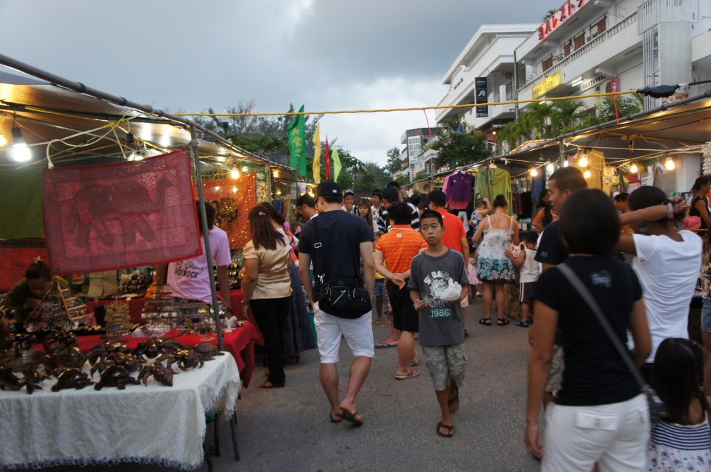 A bustling outdoor market scene with numerous people walking and browsing various stalls. The stalls are covered with canopies and are adorned with colorful fabrics, clothing, and other items for sale. The market is lit with hanging lights, and the sky above is overcast. In the background, there are buildings with signs and greenery. The atmosphere appears lively and crowded.