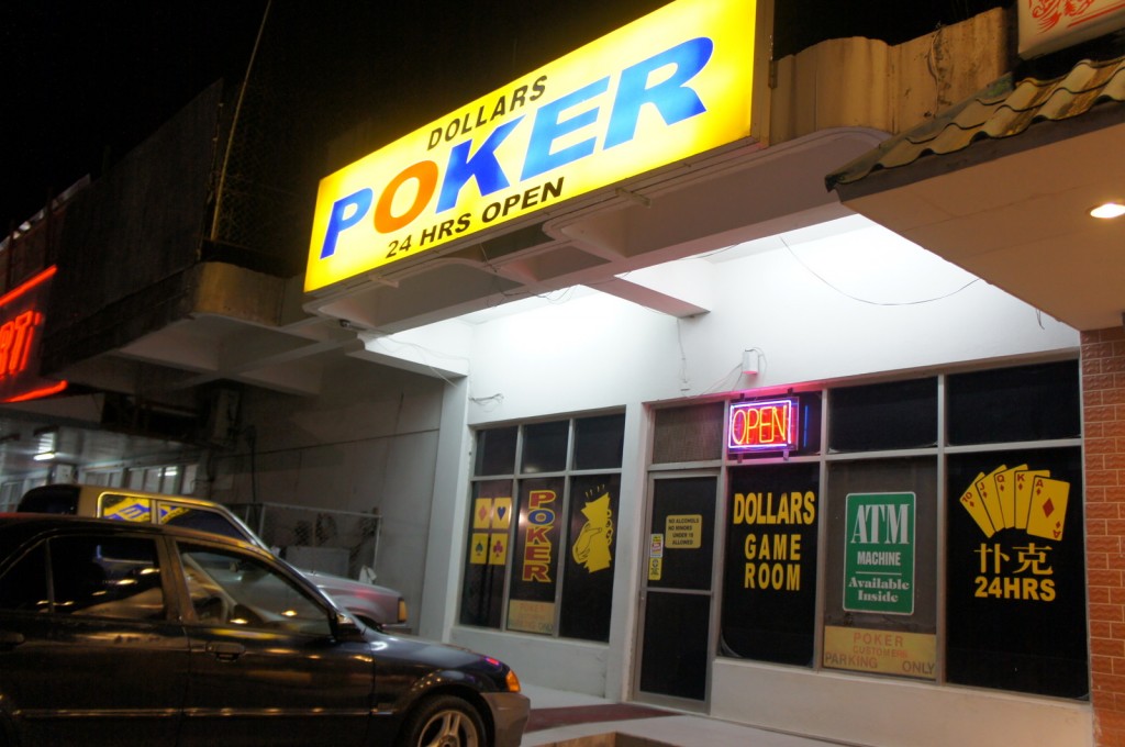 The image shows the exterior of a poker game room establishment at night. The building has a bright yellow and blue sign that reads "DOLLARS POKER 24 HRS OPEN." Below the sign, there is a neon "OPEN" sign in the window. The windows display various signs, including "DOLLARS GAME ROOM," "ATM MACHINE Available Inside," and an illustration of playing cards with the text "24HRS." A car is parked in front of the establishment. The surrounding area appears to be part of a commercial strip with other businesses.