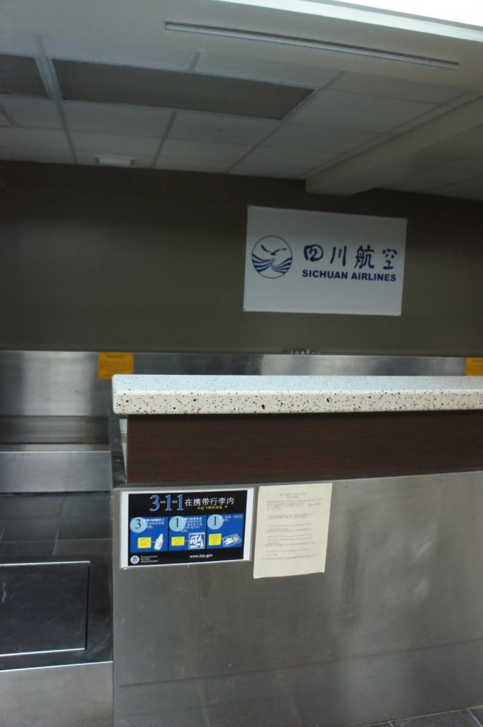 The image shows a check-in counter for Sichuan Airlines. The counter has a speckled countertop and a sign above it with the Sichuan Airlines logo and name in both Chinese and English. There is a poster on the front of the counter with information in Chinese, and another notice or document is attached next to it. The background includes a ceiling with fluorescent lights and a metallic wall behind the counter.
