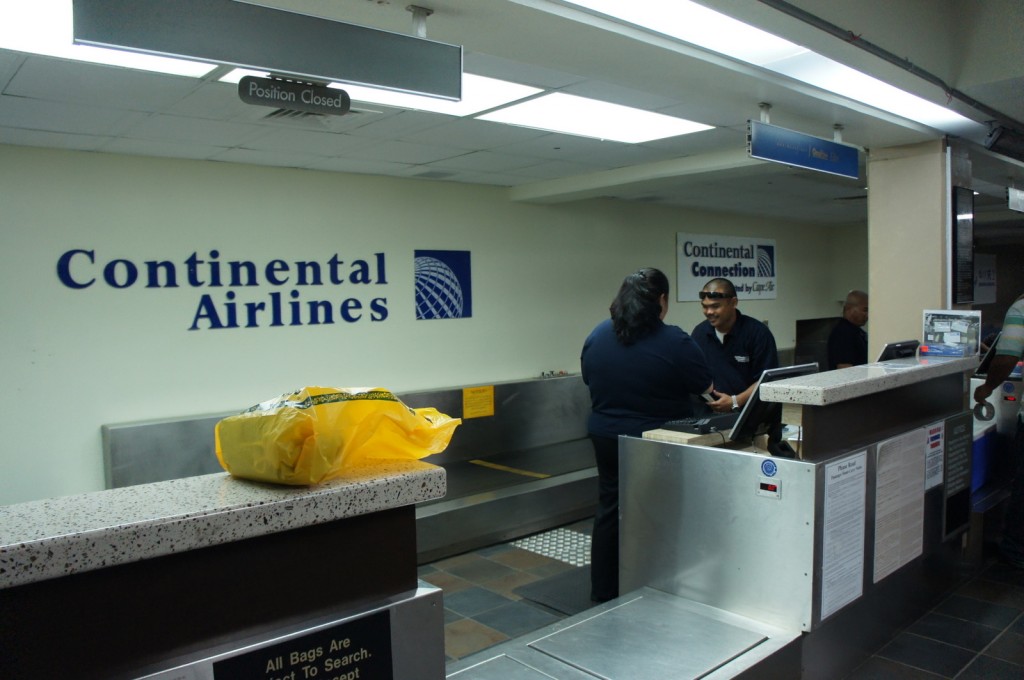 The image shows a Continental Airlines check-in counter at an airport. There are three airline staff members behind the counter, with one of them interacting with a customer. The counter has a sign above it that reads "Position Closed." There is a yellow bag on the counter in the foreground. The wall behind the counter displays the Continental Airlines logo and a sign for "Continental Connection." The area appears to be part of an airport terminal.