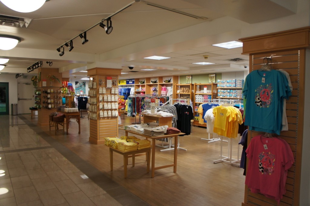 The image shows the interior of a gift shop or souvenir store. The store is well-lit and organized, with various items on display. There are racks of colorful t-shirts, shelves with neatly arranged merchandise, and display stands with smaller items. The floor is tiled, and the ceiling has track lighting. The overall atmosphere is clean and inviting, with a variety of products available for purchase.