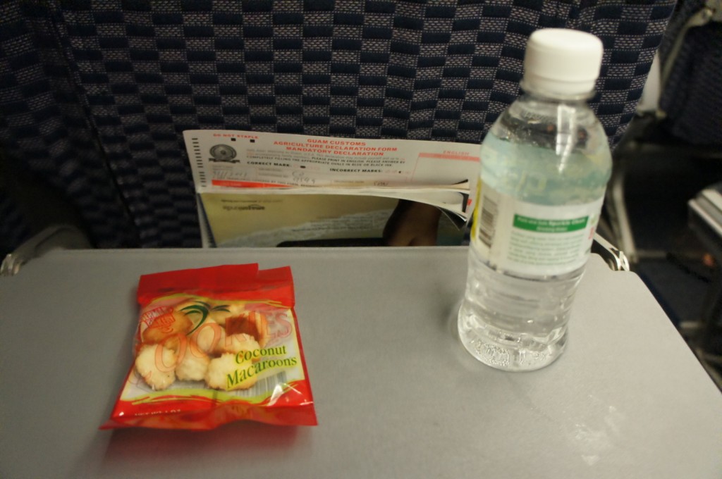 The image shows an airplane tray table with a small bottle of water and a packet of coconut macaroons on it. In the background, there is the back of an airplane seat with a pocket containing a customs declaration form and other documents.