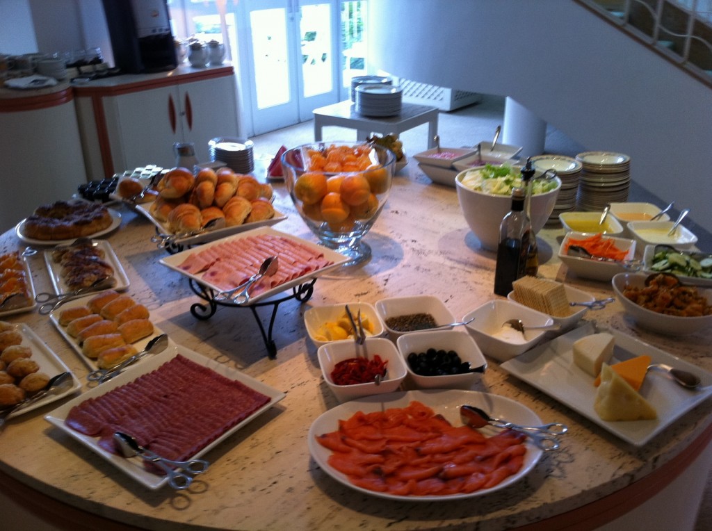 The image shows a buffet table set with a variety of food items. There are plates of sliced meats, cheeses, and smoked salmon. Bowls of fresh vegetables, olives, and condiments are also present. Additionally, there are trays of pastries, croissants, and bread rolls. A large bowl of oranges is placed in the center of the table. The table is situated in a well-lit room with a counter and a coffee machine in the background. Plates and utensils are neatly stacked and ready for use.