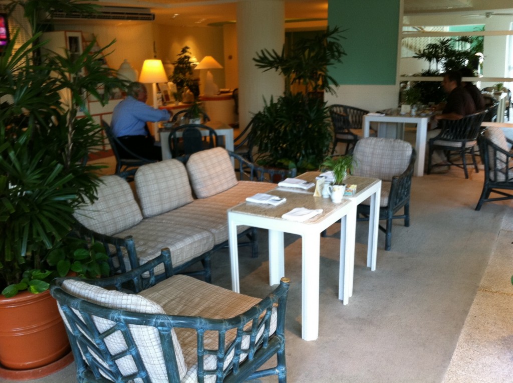 The image shows a cozy indoor seating area, likely in a caf&eacute; or restaurant. The space is furnished with wicker chairs and cushioned sofas arranged around white tables. The tables are set with napkins, small potted plants, and other dining essentials. The area is decorated with numerous green plants, creating a lush and inviting atmosphere. In the background, a few people are seated at tables, engaged in conversation or dining. The lighting is warm, with table lamps adding to the ambiance.