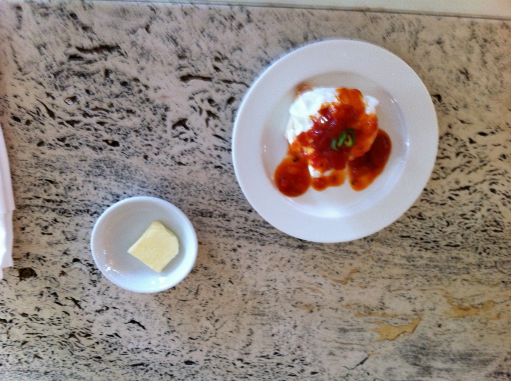 The image shows a white plate with a serving of food topped with a red sauce and garnished with a green herb, placed on a marbled surface. Next to it, there is a small white bowl containing a piece of butter.