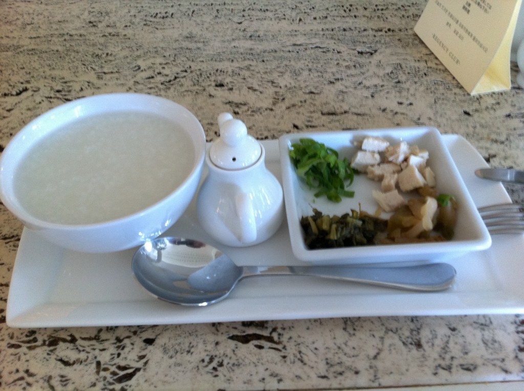 The image shows a meal consisting of a bowl of white rice porridge, a small white teapot, and a rectangular plate with three compartments containing chopped green onions, diced chicken, and pickled vegetables. A spoon and a fork are placed on the tray next to the plate. The meal is served on a white tray on a textured surface.