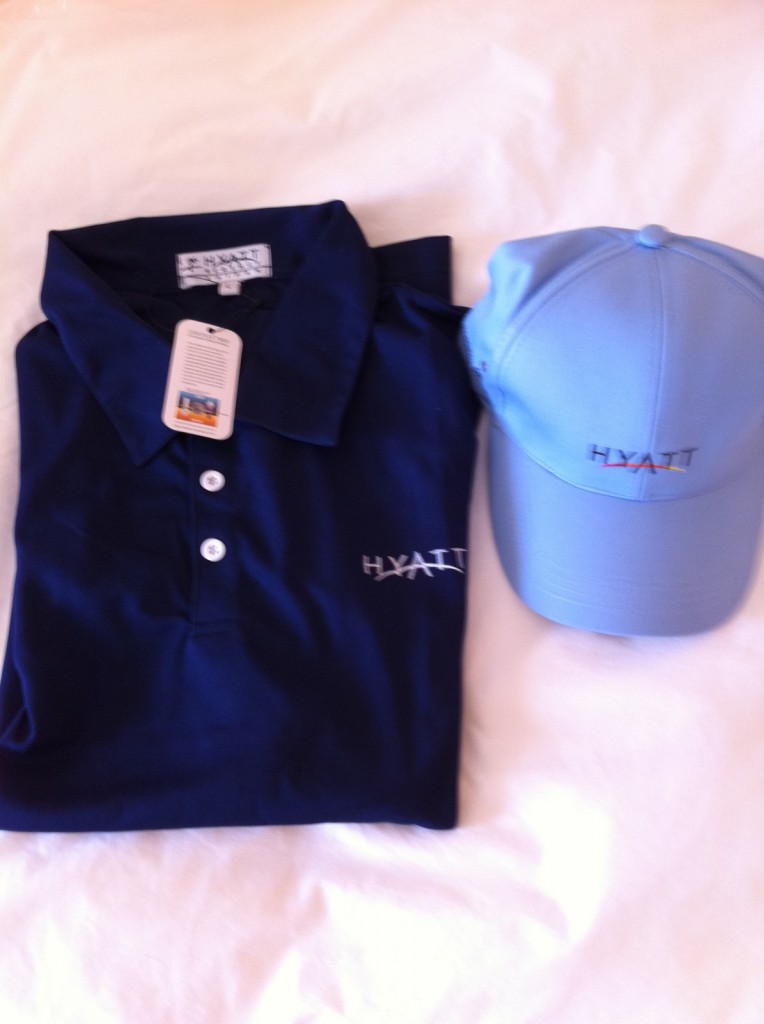 The image shows a folded navy blue polo shirt and a light blue baseball cap placed on a white surface. Both the shirt and the cap have the "Hyatt" logo embroidered on them. The polo shirt has a tag attached to it.