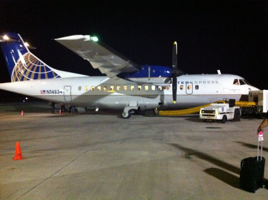 A small commercial airplane is parked on the tarmac at night. The aircraft has a blue and white color scheme with a logo on the tail and the text "UNITED EXPRESS" on the fuselage. The registration number "N14834" is visible on the side. There are orange cones placed around the plane, and a ground service vehicle is positioned near the aircraft. A person with a suitcase is visible in the foreground on the right side of the image.