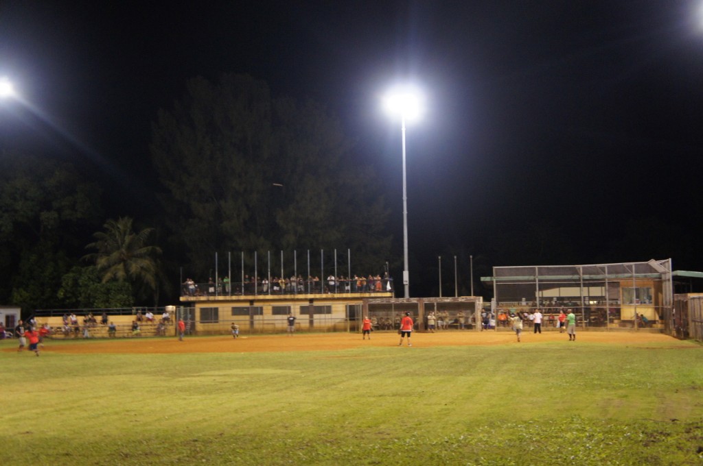 A nighttime scene of a baseball or softball game in progress. The field is illuminated by bright floodlights, and players are visible on the field. In the background, there are spectators seated in bleachers, and some people are standing. The area is surrounded by trees, and there is a building or structure behind the bleachers. The grass on the field is green, and the infield appears to be made of dirt.