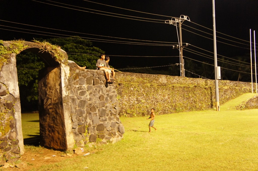 The image shows a nighttime scene with a stone wall and an archway. Two people are sitting on top of the wall, while a child is standing on the grass below, looking up at them. The area is illuminated by streetlights, and there are power lines visible in the background. The wall appears to be old and has some vegetation growing on it. The scene has a relaxed and casual atmosphere.
