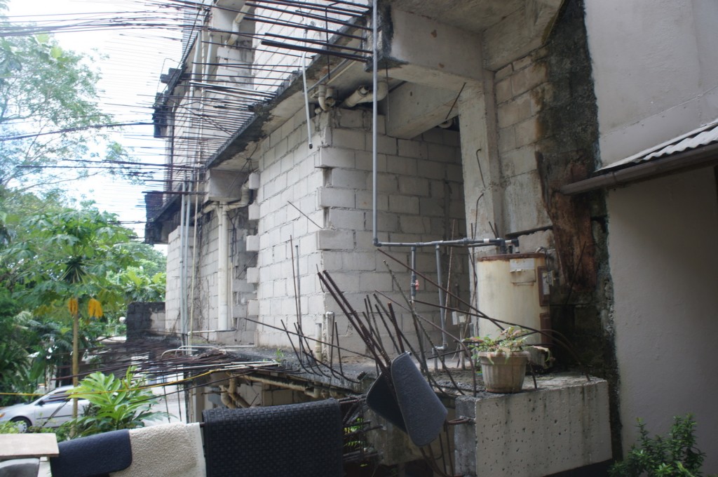 The image shows an unfinished or abandoned building with exposed concrete blocks and rebar protruding from the structure. There are various pipes and a water heater attached to the building. In the foreground, there are some chairs and a small potted plant on a concrete ledge. The surrounding area has lush green vegetation, including trees and plants. The overall scene appears to be in a state of disrepair or construction.