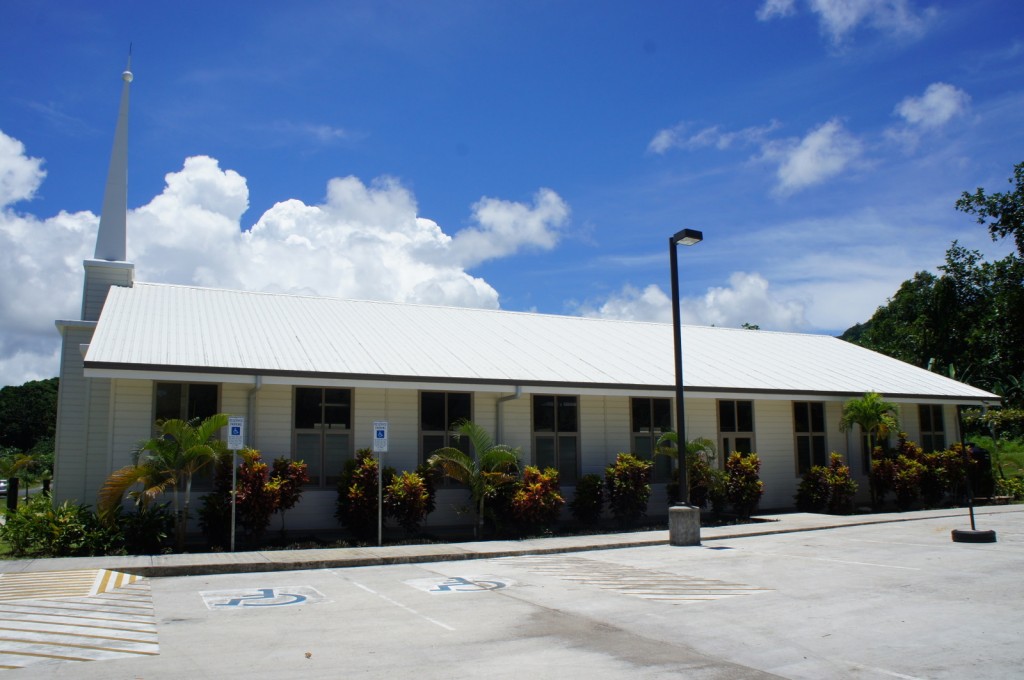The image shows a single-story building with a white exterior and a steeply pitched roof, topped with a tall steeple. The building is surrounded by well-maintained landscaping, including various shrubs and small palm trees. In front of the building, there is a parking lot with clearly marked handicapped parking spaces. The sky above is bright blue with scattered white clouds, indicating a sunny day.