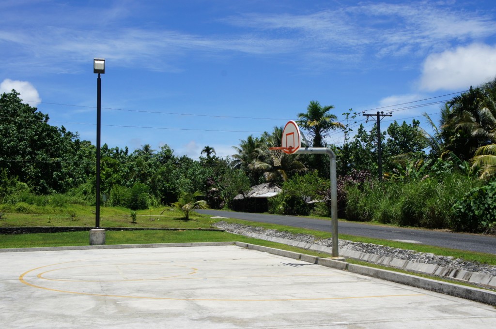 The image shows an outdoor basketball court with a single basketball hoop. The court is situated near a road and is surrounded by lush green vegetation, including trees and bushes. There is a tall streetlight near the court, and the sky is clear with a few clouds, indicating a sunny day.