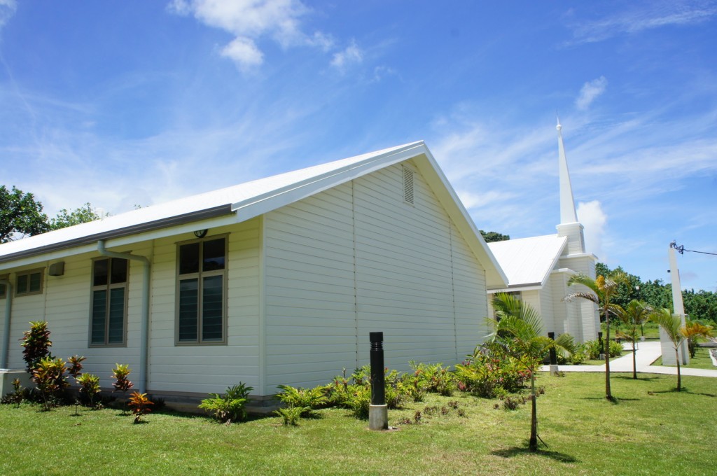 The image shows a white, single-story building with a steeply pitched roof and a tall steeple, suggesting it is a church. The building is surrounded by a well-maintained lawn with small plants and shrubs. The sky is clear with a few clouds, indicating a sunny day.