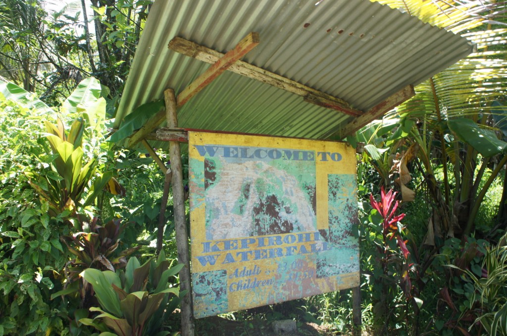 The image shows a weathered sign under a corrugated metal roof, surrounded by lush tropical vegetation. The sign reads "Welcome to Kepirohi Waterfall" and includes information about admission prices for adults and children. The sign appears to be old and faded, with some parts difficult to read. The area around the sign is dense with green plants and trees, indicating a natural, outdoor setting.