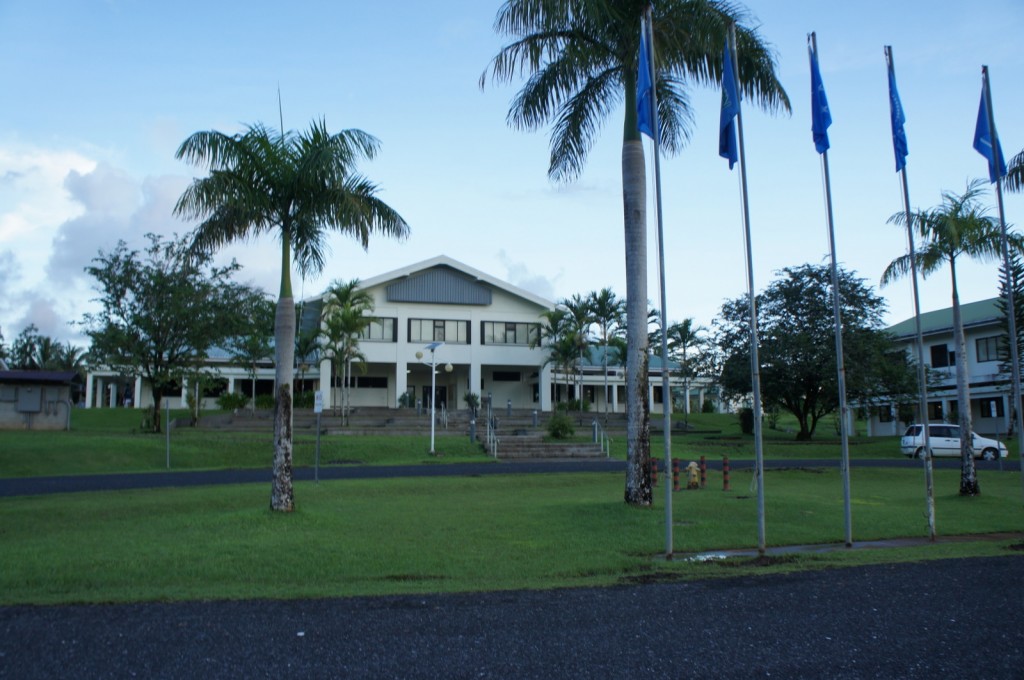The image shows a large, two-story building with a gabled roof, surrounded by a well-maintained lawn and several palm trees. In front of the building, there are multiple flagpoles with blue flags. The building appears to be institutional, possibly a school or government building. There is a paved pathway leading up to the entrance, and a white vehicle is parked to the right side of the image. The sky is clear with some clouds.