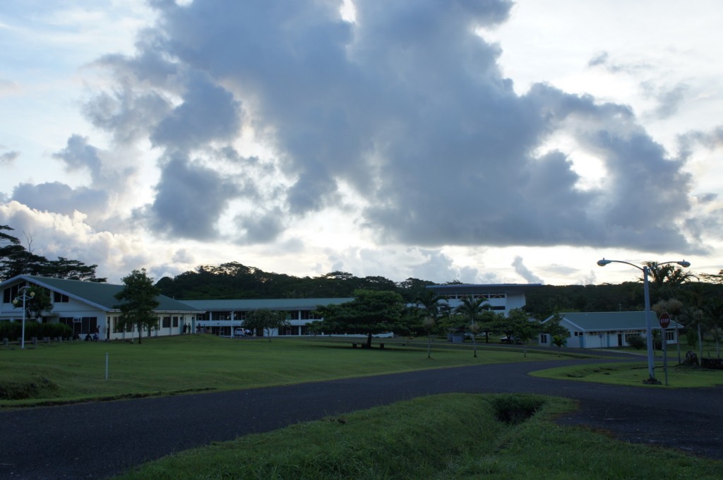 The image shows a serene landscape with a few buildings that have green roofs, likely part of a campus or residential area. The buildings are surrounded by well-maintained grassy areas and trees. A paved road curves through the scene, and there are streetlights and a stop sign visible. The sky is filled with large, dramatic clouds, suggesting an overcast or partly cloudy day. The overall atmosphere is calm and peaceful.
