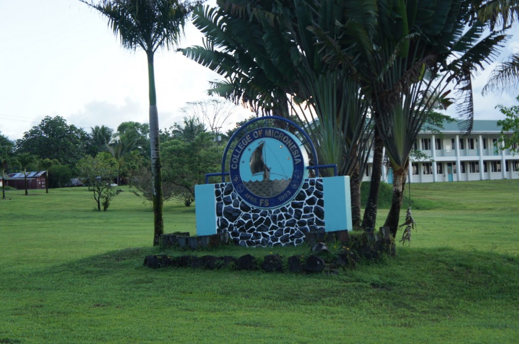 The image shows a sign for the College of Micronesia, situated on a grassy lawn with palm trees and other greenery in the background. The sign features a circular emblem with text and an image of a person, and it is mounted on a stone base. In the background, there is a multi-story building with a green roof and multiple windows. The overall setting appears to be a tropical or subtropical campus environment.