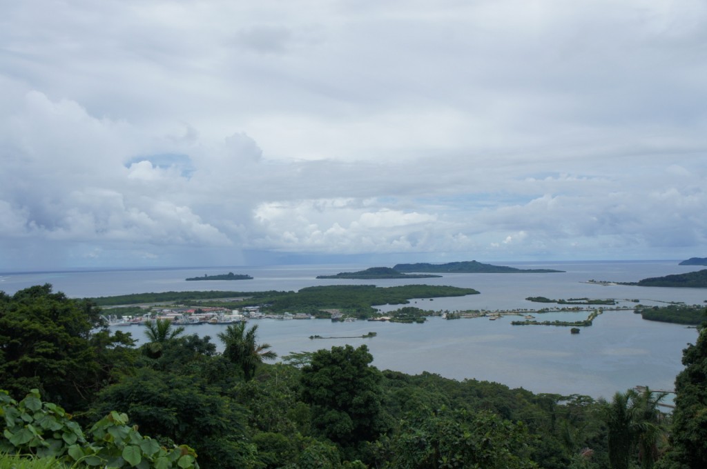 A scenic view of a coastal area with lush green vegetation in the foreground. The image shows several small islands and peninsulas surrounded by calm, blue water. The sky is mostly cloudy with patches of blue peeking through. The overall atmosphere is serene and natural.