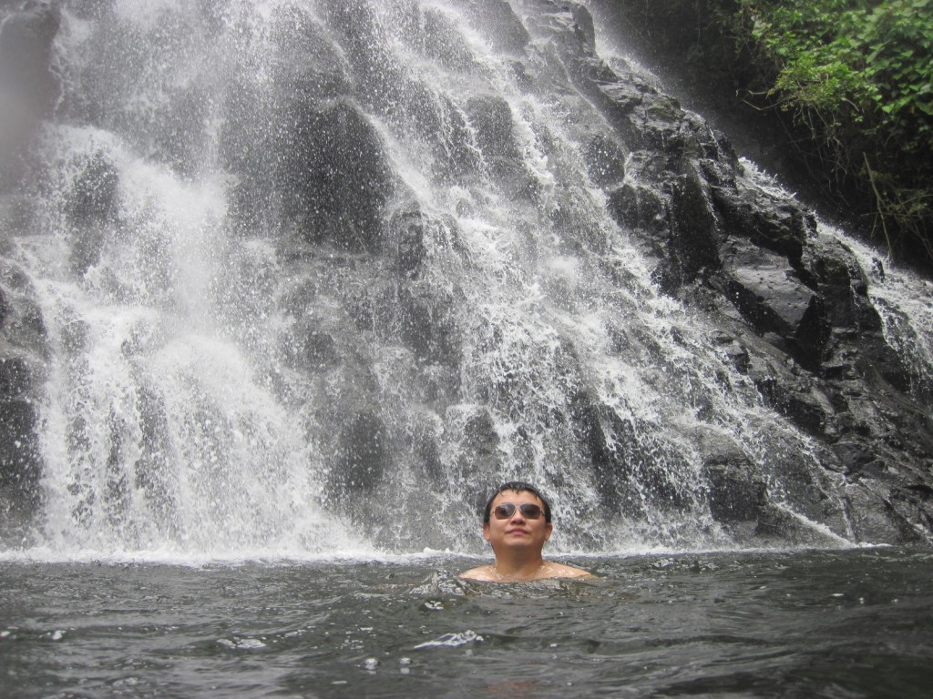 A person wearing sunglasses is partially submerged in water at the base of a large, cascading waterfall. The waterfall flows over dark, rocky terrain, and there is greenery visible on the right side of the image. The person appears to be enjoying the natural setting.